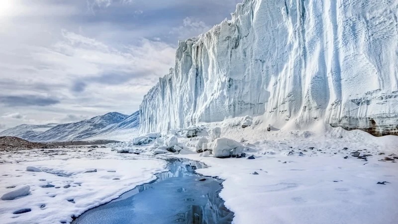 Massive ice cliff beside a snowy plain with a narrow blue meltwater stream and distant mountains