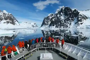 Passengers in orange jackets on a ship bow viewing snow-covered peaks mirrored in polar waters