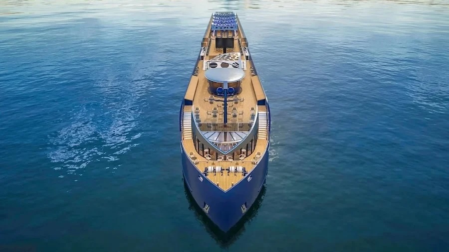 Bow-on aerial view of a large luxury yacht with teak decks, lounge seating, and calm blue sea.