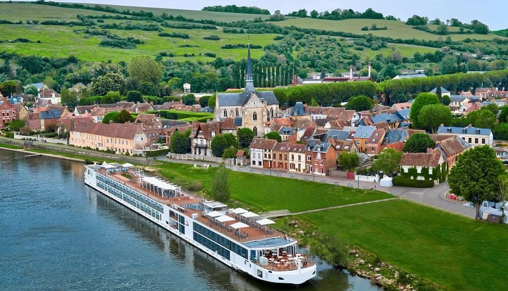 White river cruise ship docked beside a quaint village with church steeple and rolling green hills