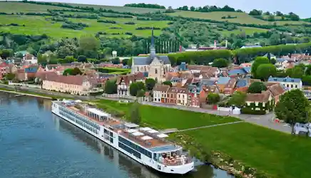 White river cruise ship docked beside a quaint village with church steeple and rolling green hills