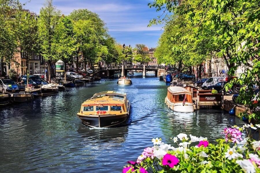 Tree-lined canal with sightseeing boats, bridges, parked cars and flowers in foreground
