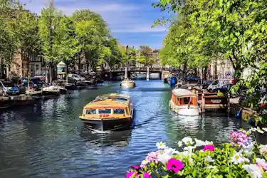 Tree-lined canal with sightseeing boats, bridges, parked cars and flowers in foreground