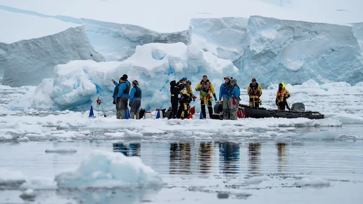 Researchers in bright cold-weather gear on an ice floe beside a rigid inflatable boat and iceberg