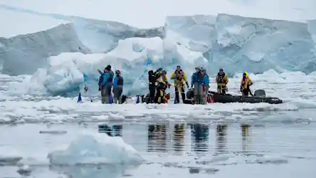Researchers in bright cold-weather gear on an ice floe beside a rigid inflatable boat and iceberg