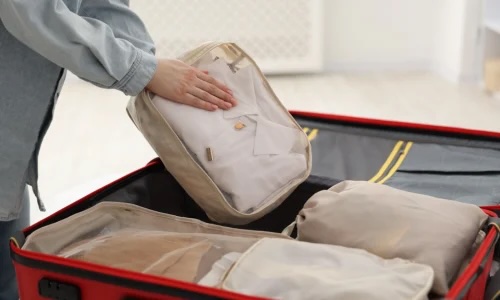 Hand placing beige packing cube with folded shirt into open red suitcase with other packing cubes