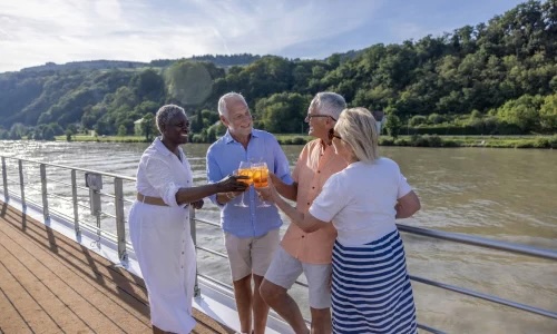 Four older adults on a riverside boardwalk toasting orange drinks, hills and river behind them