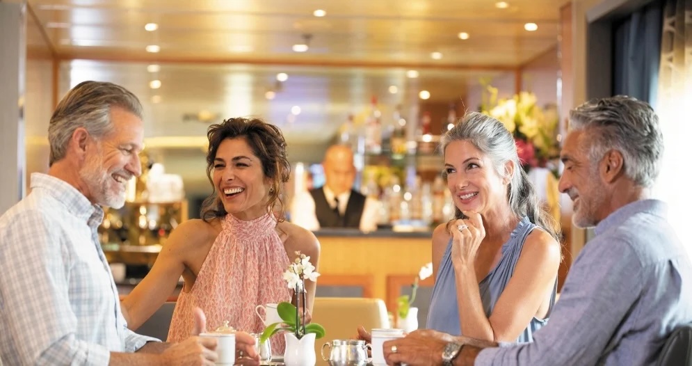 Four smiling middle-aged adults in two couples chatting and holding coffee cups in a ship lounge.