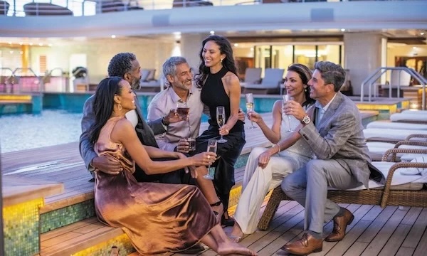Six well-dressed adults laughing and toasting by a cruise ship pool deck at dusk.