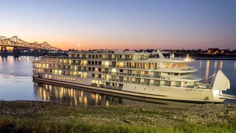 White multi-deck river cruise ship lit at dusk, moored by a riverbank with bridge and city lights