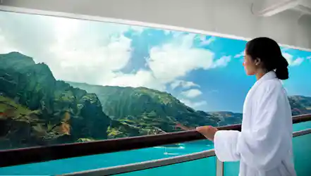 Woman in white robe on cruise balcony gazing at green cliffs and turquoise ocean.