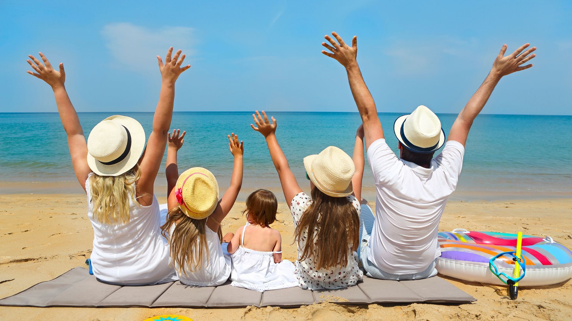 Parents and three children on a beach mat facing the ocean, arms raised, wearing sun hats