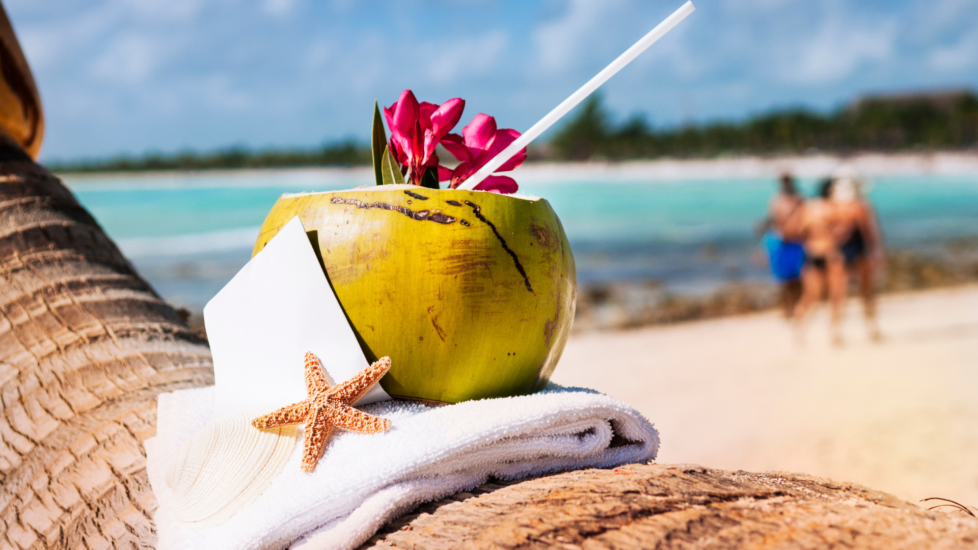 Coconut drink with straw and pink flowers on towel by turquoise sea, starfish in foreground
