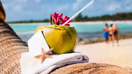 Coconut drink with straw and pink flowers on towel by turquoise sea, starfish in foreground