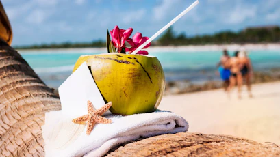Coconut drink with straw and pink flowers on towel by turquoise sea, starfish in foreground