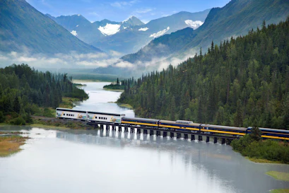 Passenger train crossing a trestle over a river in a forested valley with snow-capped peaks