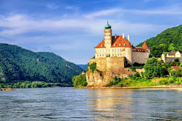 Castle with red tiled roofs and green onion dome on cliff above a river, forested hills