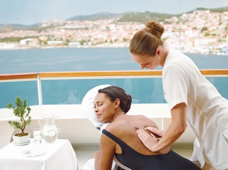Woman in black swimsuit receiving a back massage on a yacht deck with coastal town view