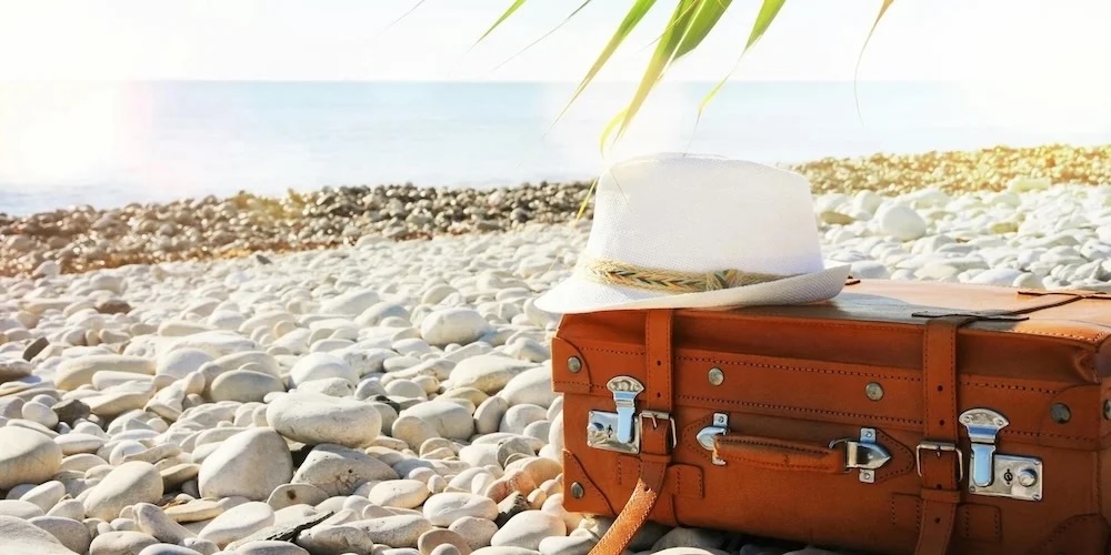 Brown leather suitcase and white straw hat on pebble beach, suggesting travel.