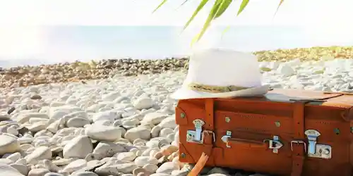 Brown leather suitcase and white straw hat on pebble beach, suggesting travel.