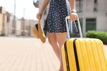 Person in floral dress pulling a yellow rolling suitcase and holding a straw hat on a city sidewalk
