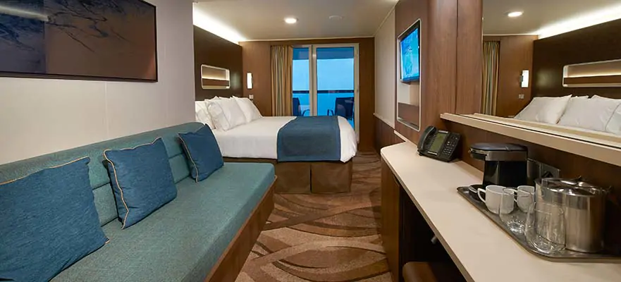 Cruise ship balcony stateroom: teal sofa with pillows, bed, desk with coffee station, ocean view.