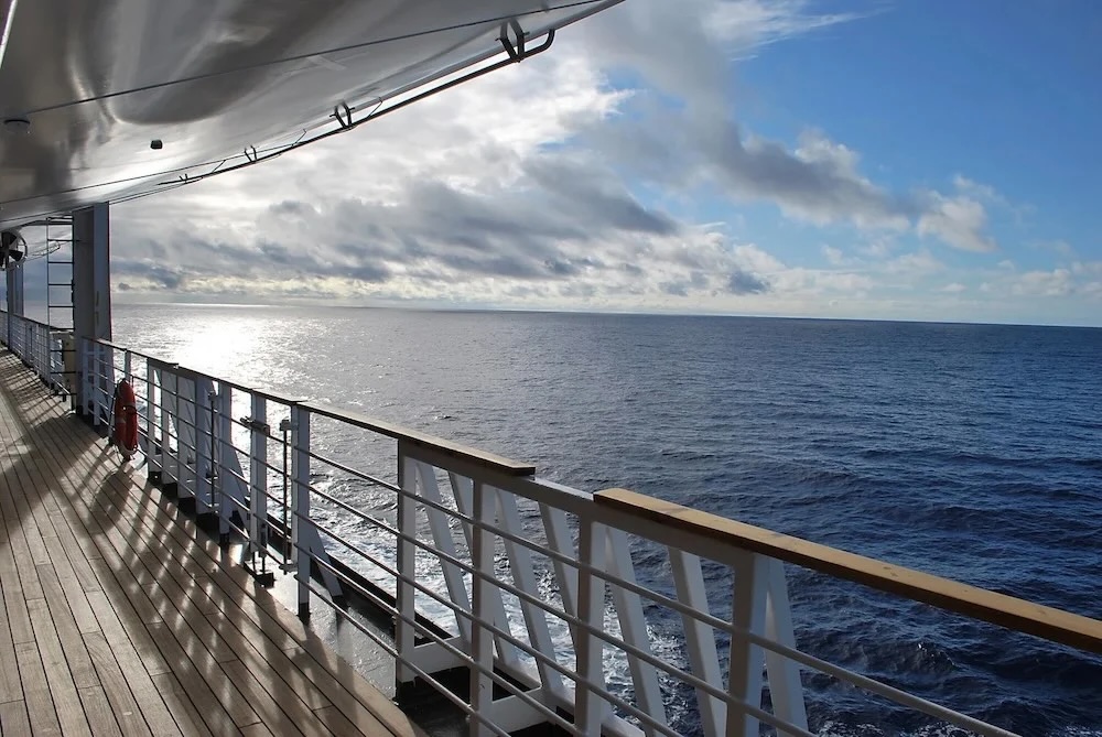 View from a cruise ship deck showing wooden railing, lifebuoy, and sunlit ocean horizon.