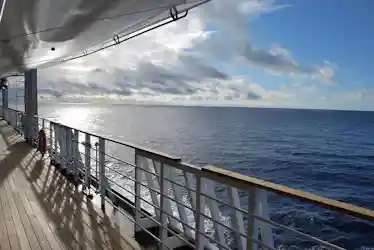 View from a cruise ship deck showing wooden railing, lifebuoy, and sunlit ocean horizon.