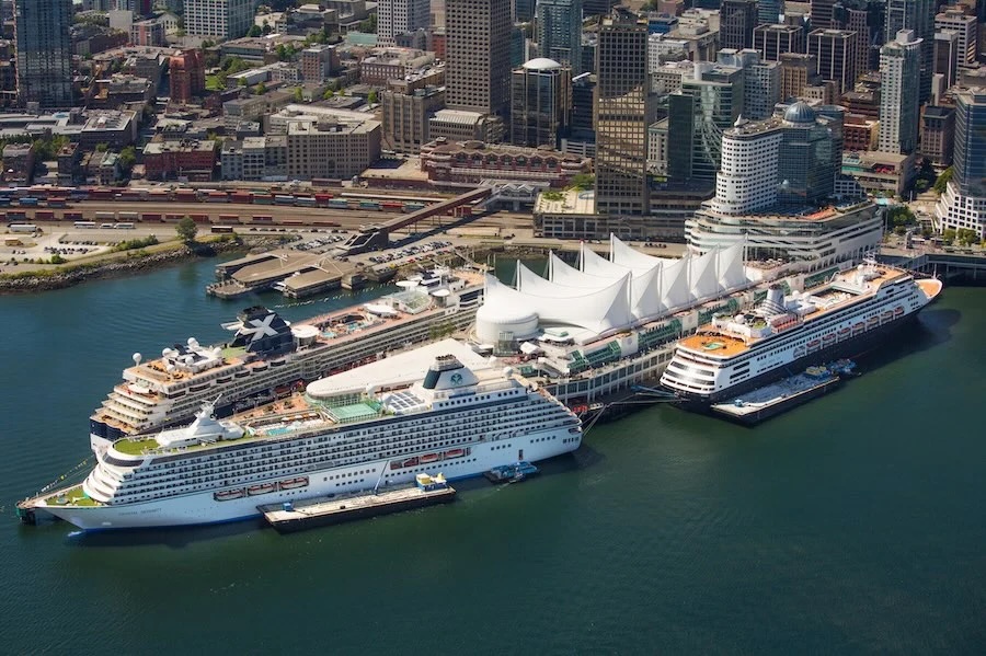 Three cruise ships docked beside a white sail-roofed waterfront terminal and city skyline