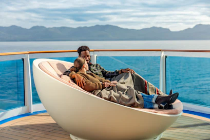 Couple cuddling under a blanket on a cruise ship deck lounger with ocean and mountains