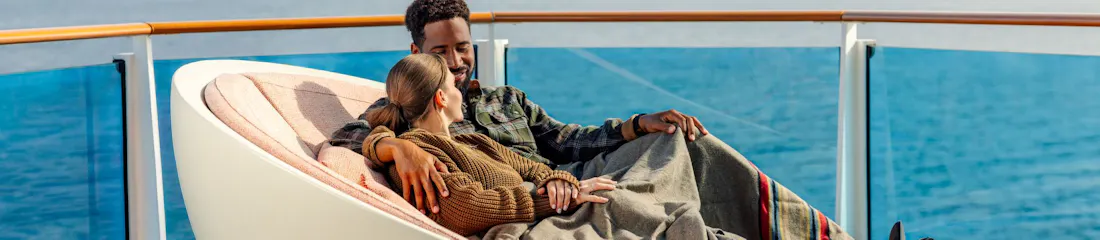 Couple cuddling under a blanket on a cruise ship deck lounger with ocean and mountains