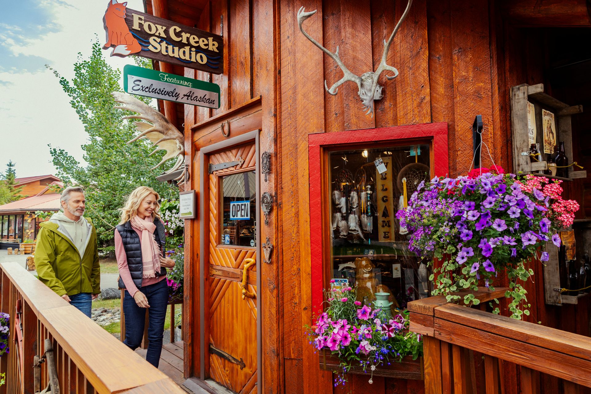 Couple walking past Fox Creek Studio wooden storefront with antlers, hanging flowers, OPEN sign