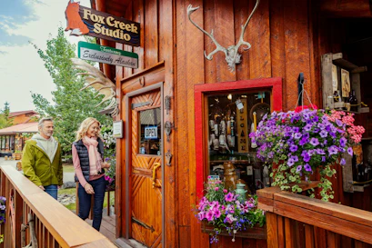 Couple walking past Fox Creek Studio wooden storefront with antlers, hanging flowers, OPEN sign