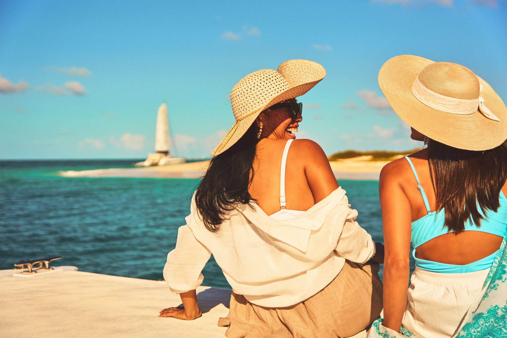 Two women in wide sun hats sitting on a dock, facing turquoise ocean with distant lighthouse