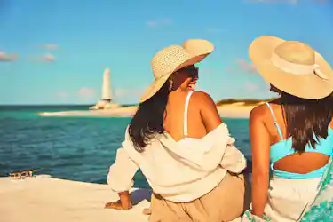 Two women in wide sun hats sitting on a dock, facing turquoise ocean with distant lighthouse