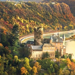 Medieval stone castle on a river bend with round tower and conical turrets amid autumn forest.