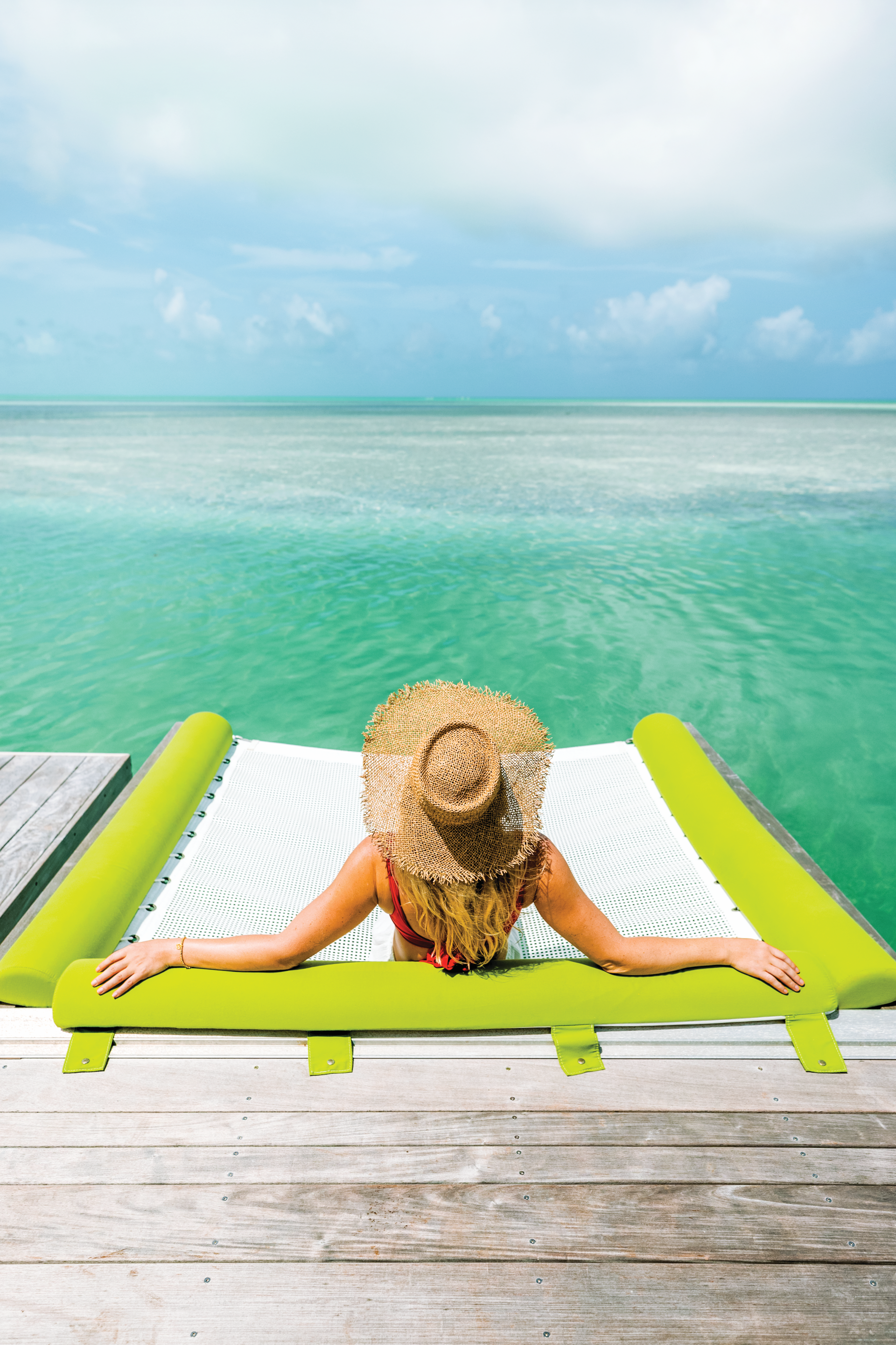 Woman in straw hat lounging on dock hammock, arms out, facing turquoise sea and sky.