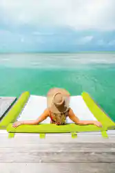 Woman in straw hat lounging on dock hammock, arms out, facing turquoise sea and sky.