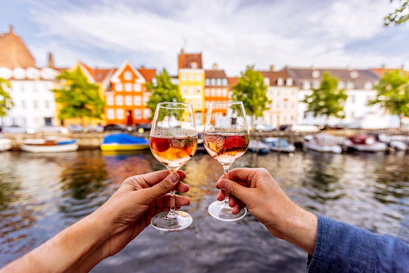 Two hands toasting rosé glasses with strawberries over a canal and colorful waterfront houses