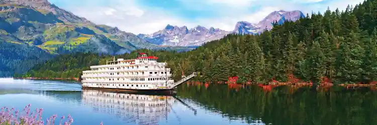 White multi-deck paddle steamer on calm mountain lake reflecting evergreens and alpine peaks