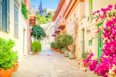 Narrow Greek lane with pastel houses, balconies, potted plants, Greek flag, pink bougainvillea