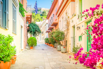 Narrow Greek lane with pastel houses, balconies, potted plants, Greek flag, pink bougainvillea