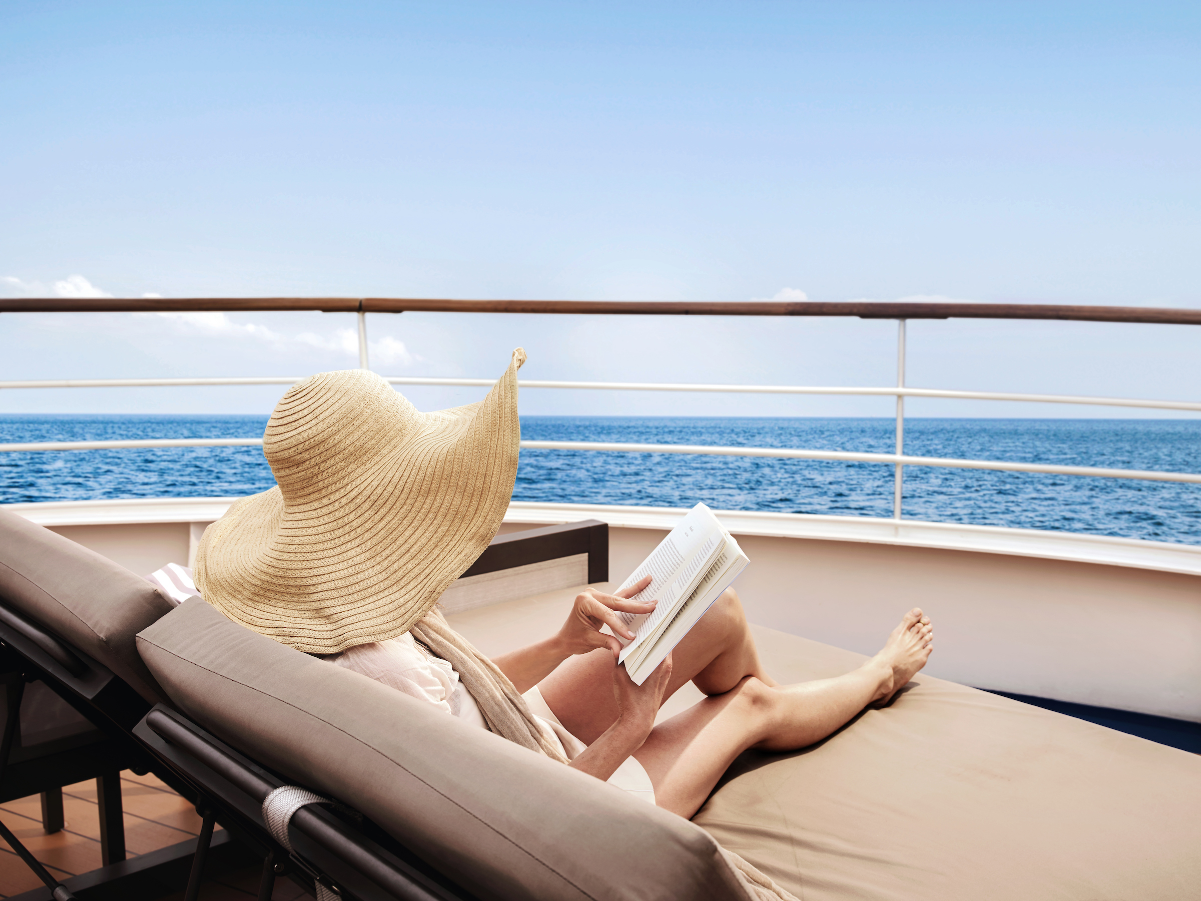 Person reclining on a yacht deck chair wearing wide sun hat and reading with ocean view.