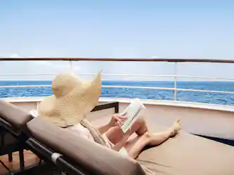 Person reclining on a yacht deck chair wearing wide sun hat and reading with ocean view.