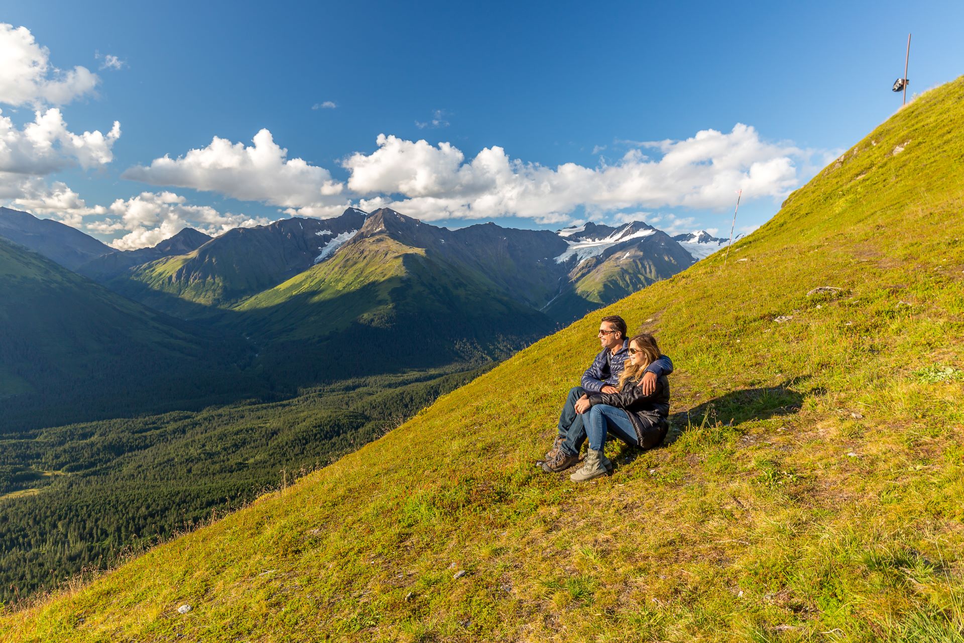 Couple seated on grassy alpine slope embracing, looking at glacier-capped mountain valley.