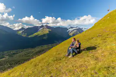 Couple seated on grassy alpine slope embracing, looking at glacier-capped mountain valley.