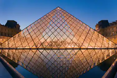 Glass Louvre Pyramid illuminated at dusk with symmetrical reflection in museum courtyard pool