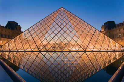 Glass Louvre Pyramid illuminated at dusk with symmetrical reflection in museum courtyard pool