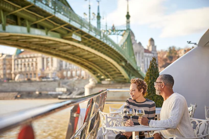 Couple smiling, holding wine at a riverside table under a green arched bridge with city backdrop.