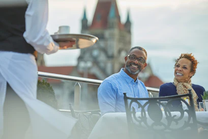 Couple smiling as a waiter brings a tray while they dine on a rooftop terrace with church spires